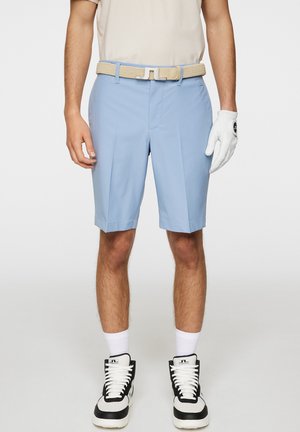 Shorts de golf bleu clair en tissu lisse, avec un design sur mesure et des plis, associés à une ceinture tissée beige. Gant blanc et baskets.