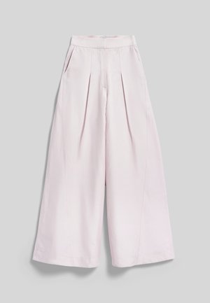 Femme aux cheveux bruns bouclés portant un blazer ceinturé rose pâle et un pantalon large assorti sur un fond gris clair.