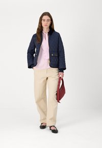 Navy quilted jacket with a zip front and gold hardware, paired with a light pink ruffled blouse and beige wide-leg trousers, holding a red bag.