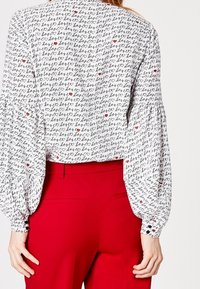 Biała bluzka z czarnym wzorem tekstowym, z czerwonymi akcentami. Bufiaste rękawy z marszczonymi mankietami, w połączeniu z czerwonymi spodniami.