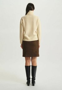 Pull crème avec col montant, associé à une jupe marron texturée. Le mannequin porte des bottes noires jusqu'aux genoux. Vue de dos.