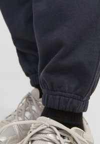 Mørkeblå joggers i blødt bomuld med elastiske manchetter, parret med lysegrå sneakers og sorte ankelstrømper. Detalje på manchetterne synlig.