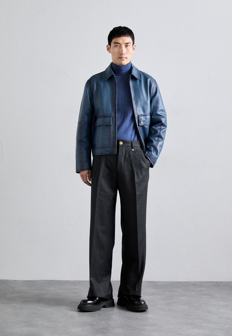 Giubbotto di pelle blu navy, abbinato a un dolcevita grigio scuro e pantaloni tagliati. Scarpe nere spesse completano l'outfit.