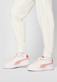 Rózsaszín és krém színű sneaker, texturált felső résszel, kerek orral, vastag fehér talppal, és korall színű részlettel a saroknál. Krém színű melegítőnadrággal párosítva.
