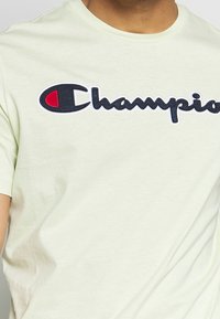 Svetle zelené bavlněné tričko s velkým vyšitým logem "Champion" v černé a červené barvě, s měkkou texturou a uvolněným střihem.