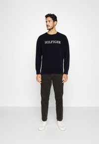 Maglione girocollo blu navy con il logo "HILFIGER" in filo chiaro. Abbinato a pantaloni scuri e sneakers bianche, che mostrano un look casual.