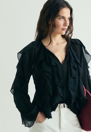 Mujer con blusa negra con volantes y pantalones anchos blancos, sosteniendo un bolso tote texturizado de color rojo oscuro, de pie frente a un fondo liso y claro.