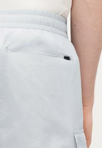 Ανοιχτό γκρι παντελόνι με ελαστική μέση και μικρή τσέπη με φερμουάρ στο πλάι, που φοριέται από ένα άτομο με ορατό χέρι.