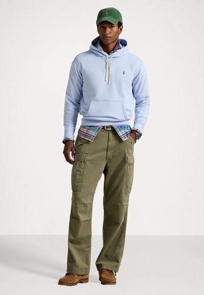 Helles blaues Kapuzenpullover mit Kordeln, über einem karierten Hemd getragen, kombiniert mit grünen Cargo-Hosen und braunen Schuhen. Es trägt eine grüne Mütze.