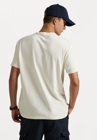 T-shirt curta de cor bege claro com um corte descontraído, textura suave e decote redondo, combinada com calções cargo escuros e um boné azul-marinho.