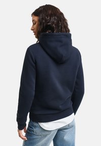 Felpa blu navy con design classico vista da dietro, cappuccio con cordino e polsini ribattuti, abbinata a jeans in denim azzurro.