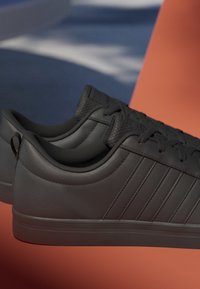 Paire de baskets basses noires avec languettes texturées et bandes latérales, présentée sur un fond géométrique bleu et orange.