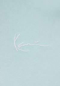 Šviesiai mėlynas audinys su lygiu paviršiumi, centre šiek tiek žemiau esančiuose vietoje siūtu baltu parašu.