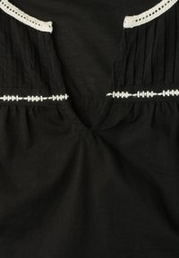 Vêtement noir avec un col en V, présentant des coutures décoratives blanches le long du col et sur la zone de la poitrine.