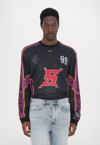 Schwarzes Langarm-Trikot mit Grafik 99, roten und lila Akzenten, Schachbrettmuster und dem Text "BRING BACK". Kombiniert mit hellblauen Jeans.