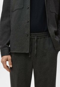 Cămașă din material textil moale, de culoare gri închis, cu două buzunare la piept și închidere cu nasturi, asortată cu pantaloni jogger cu talie elastică de aceeași culoare. Textură netedă.