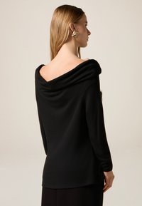 Zwarte coltrui met lange mouwen, zachte textuur en een losse pasvorm, met gedrapeerde stof bij de halslijn en een ritssluiting aan de zijkant.