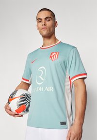 Camiseta de fútbol azul claro con acentos rojos, logo en el pecho y laterales blancos. El jugador sostiene un balón de fútbol con un patrón en blanco y negro.