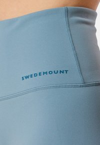 Des leggings de sport bleu clair présentent une texture lisse avec une taille élastique et le nom de la marque "SWEDEMOUNT" en bleu foncé.