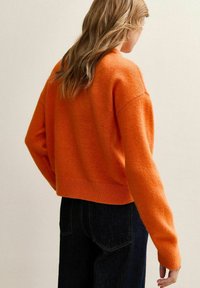 Przycięty, miękki pomarańczowy sweter z długimi rękawami, prążkowanym dolnym wykończeniem i luźnym krojem, zestawiony z ciemnoniebieskimi dżinsami.