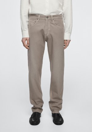 Mann trägt beige Hosen, weißes Hemd mit Knöpfen und schwarze Loafers, steht vor einem schlichten hellen Hintergrund.