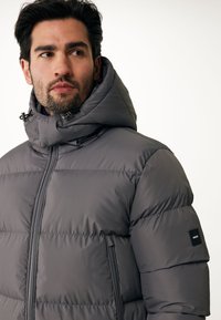 Chaqueta acolchada en gris oscuro con un alto cuello y capucha ajustable. Presenta cierre frontal y bolsillos laterales con un parche de logo en la manga.