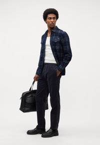 Chemise en flanelle à carreaux navy sur un débardeur blanc, associée à un pantalon en velours côtelé sombre et des mocassins noirs épais, portant un sac en cuir noir.