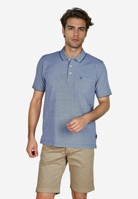 Hellblaues Poloshirt aus weichem Stoff, mit einer Knopfleiste mit drei Knöpfen, kurzen Ärmeln und einer Brusttasche. Kombiniert mit beigen Shorts.