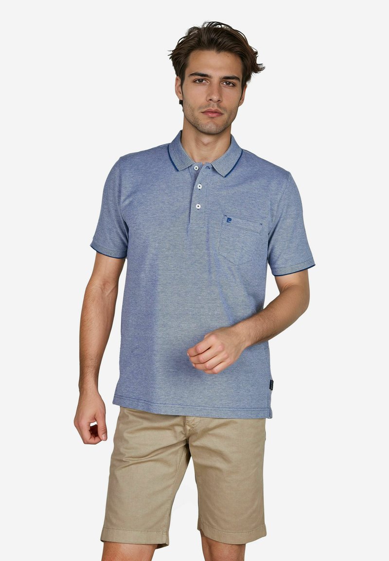 Hellblaues Poloshirt aus weichem Stoff, mit einer Knopfleiste mit drei Knöpfen, kurzen Ärmeln und einer Brusttasche. Kombiniert mit beigen Shorts.