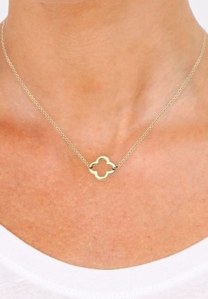 Collier délicat en chaîne d'or avec un petit pendentif en forme de quadrilobe porté sur un cou nu au-dessus d'un haut blanc.