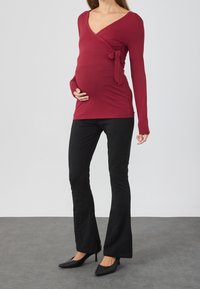 Kobieta w ciąży ubrana w buraczaną bluzkę z długim rękawem, czarne spodnie i czarne szpilki, stojąca na jednolitym tle.