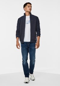 Chaqueta navy con cremallera y panel superior texturizado, combinada con una camiseta gráfica blanca, jeans azules y zapatillas blancas. Corte informal, diseño minimalista.