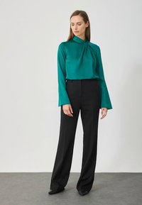 Satenasta bluza v barvi modrozelene z vozlom na izrezu vratu, kombinirana z visokim pasom črnimi hlačami. Gladek tekstil, dolgi rokavi in krojen kroj.