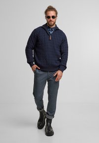 Maglione blu navy a maglia con un design a cavo testurizzato, colletto con zip, abbinato a pantaloni grigi e stivali neri alla caviglia.