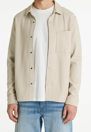 Mand iført beige knap-op tekstureret jakke over hvid t-shirt og blå jeans, stående mod en ensfarvet baggrund.