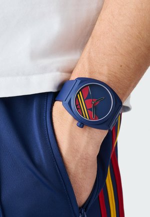 Ceas de mână albastru cu logo Adidas roșu și galben pe încheietura unei persoane care poartă pantaloni de trening bleumarin cu dungi roșii și galbene și cămașă albă.