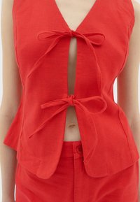 Red linen top featuring a deep V-neck and front ties, with a slightly flared hem. The fabric has a textured finish. Matching red pants visible.