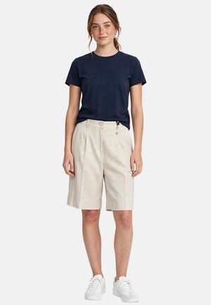 Giovane donna che indossa una t-shirt blu navy, pantaloncini beige fino al ginocchio e scarpe da ginnastica bianche, in piedi davanti a uno sfondo semplice.