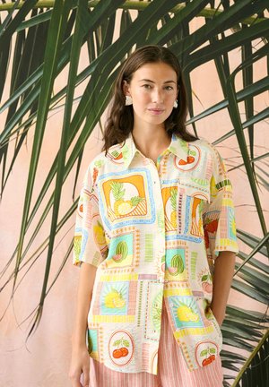 Hosszú barna hajú nő színes, gyümölcsmintás rövid ujjú inget és fehér fülbevalókat visel, nagy zöld pálmalevelek előtt áll.