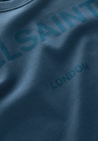 Sudadera color teal con una textura suave, que presenta letras en negrita y ligeramente en relieve que dicen "ELSAINT" y "LONDRES" en un tono más oscuro de teal.