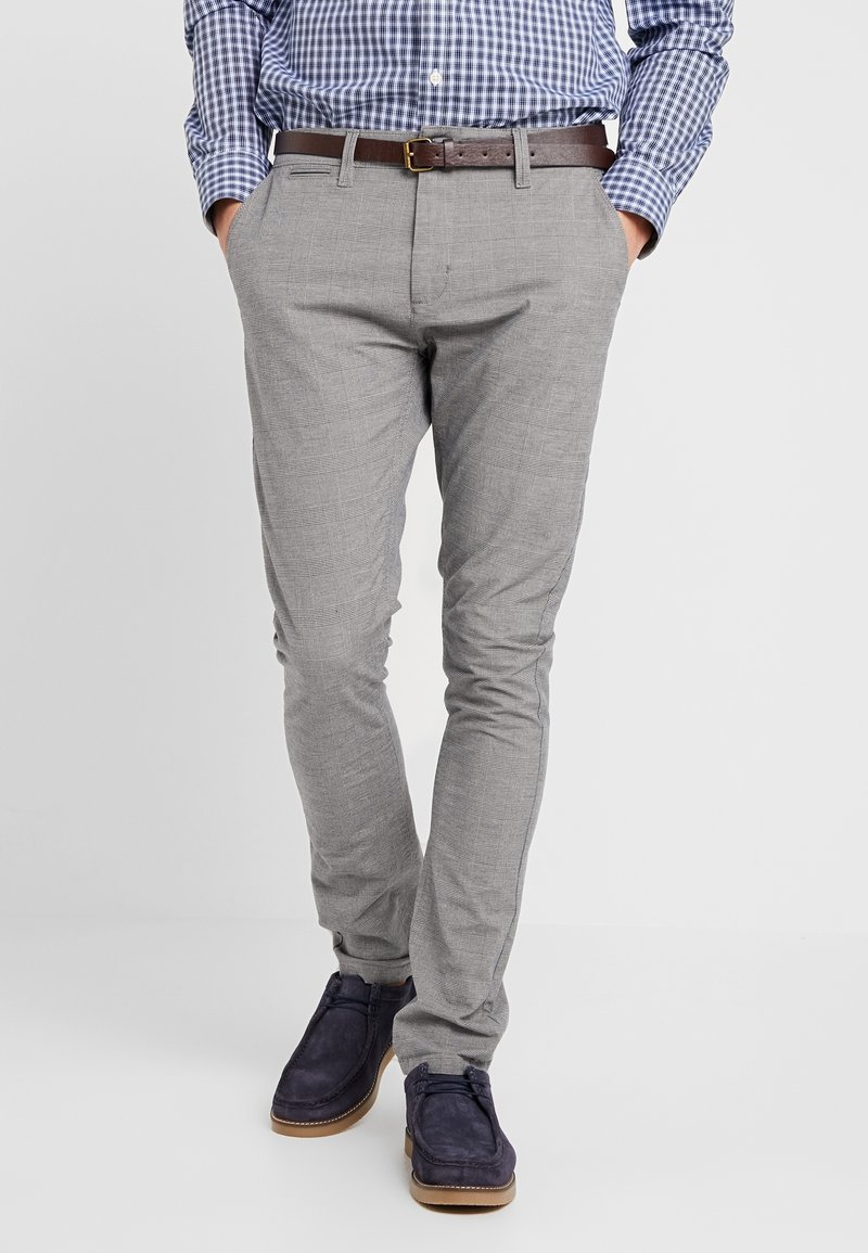 Homme portant un pantalon à carreaux gris clair, une ceinture en cuir marron, une chemise à carreaux bleue et des chaussures en daim bleu marine, debout les mains dans les poches.