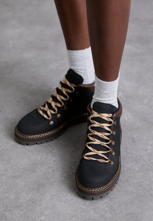 Botas de senderismo de ante negro con cordones beige y rojos, ojales de metal y acentos marrones. Suela texturizada con patrones de banda de rodadura visibles.