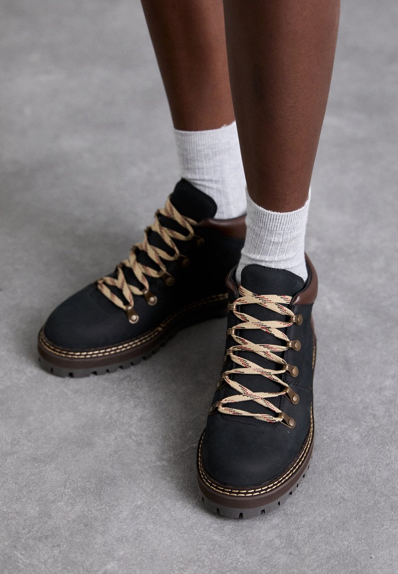 Melnas zamšādas pārgājienu zābaki ar bēšiem un sarkanim auklām, metāla acīm un brūniem akcentiem. Strukturēta zole ar redzamām protektora zīmēm.