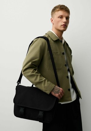 Young man wearing an olive green jacket and white shirt holds a black messenger bag over one shoulder, showing a detailed hand tattoo.