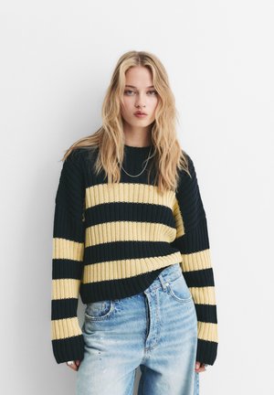 Young woman with long blonde hair wearing a black and yellow striped knit sweater and light blue high-waisted jeans against a white background.