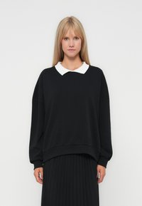 Svart hettegenser med hvit krage, oversized passform, ribbede mansjetter og kant, båret over et plissert svart skjørt. Glatt tekstur, uformell design.