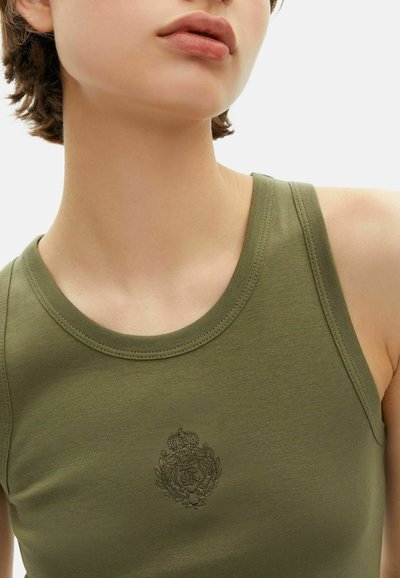 Close-up van een persoon die een olijfgroene mouwloze top draagt met een klein geborduurd wapen op de borst.