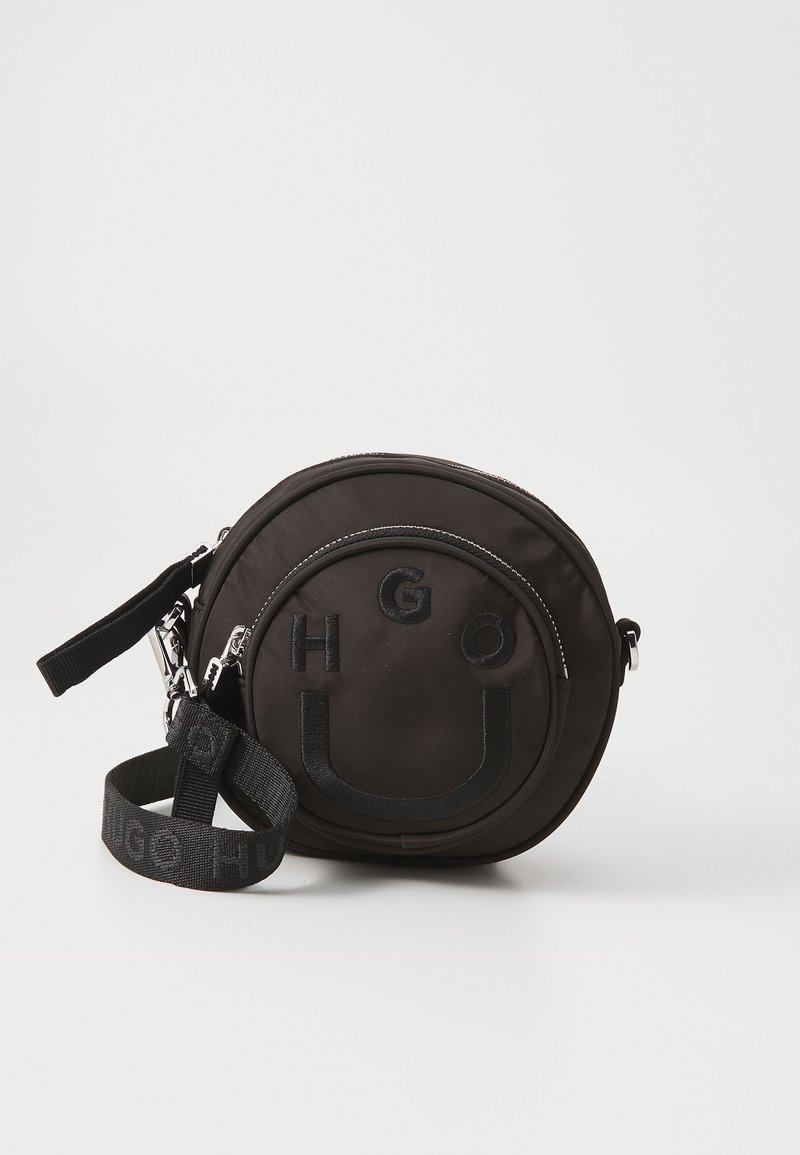 Black circular bag made of smooth fabric, featuring embossed "HUGO" logo, zip closure, and detachable strap with logo detail.
