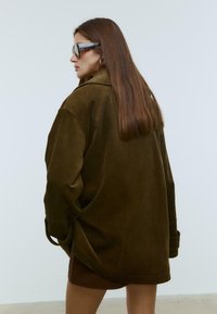Braune Cordjacke im Oversized-Stil mit Kragen-design, Seitentaschen und hochgekrempelten Ärmeln. Das Model trägt zudem eine Sonnenbrille und hat glattes Haar.