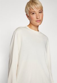 Langermet, elfenbensfarget topp laget av myk stoff, med rund hals og litt løs passform. En subtil "BORG" logo er plassert på brystet.